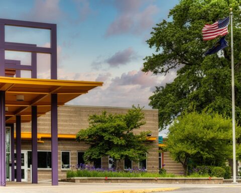 A flag pole with an American flag sits in front of a school building in the day.
