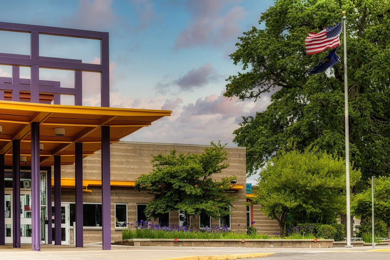 A flag pole with an American flag sits in front of a school building in the day.