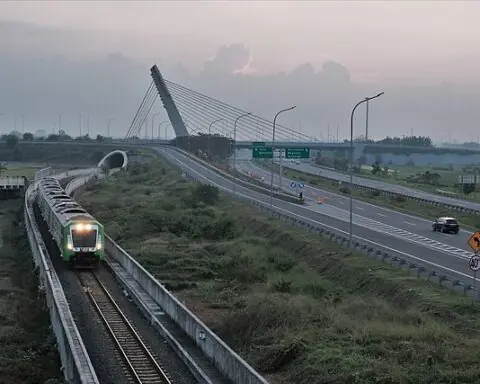 an aerial view shows a roll road parallel to a train coming down tracks in front of a cloudy sky