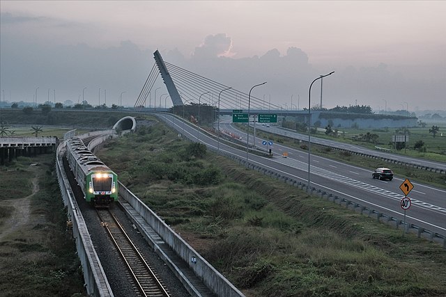 an aerial view shows a roll road parallel to a train coming down tracks in front of a cloudy sky