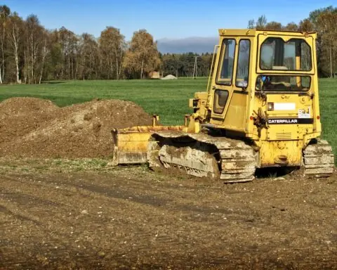 An excavator pushes dirt into a pile in front of a grassy field and trees.