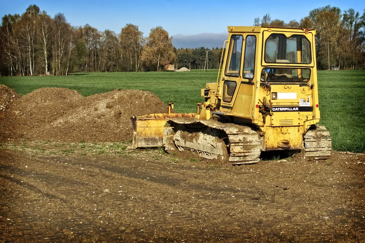 An excavator pushes dirt into a pile in front of a grassy field and trees.