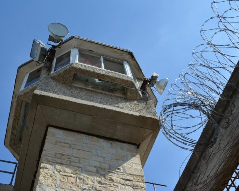 A guard tower looms over a barbed wire fence and other security measures.