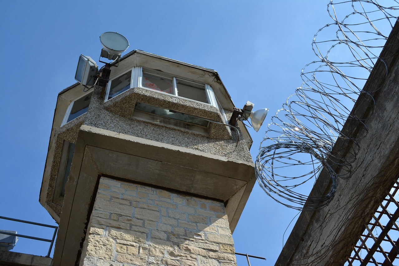 A guard tower looms over a barbed wire fence and other security measures.