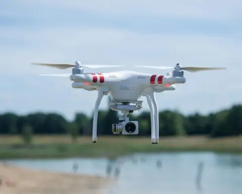 A drone is pictured above a small body of water and trees in the distance.