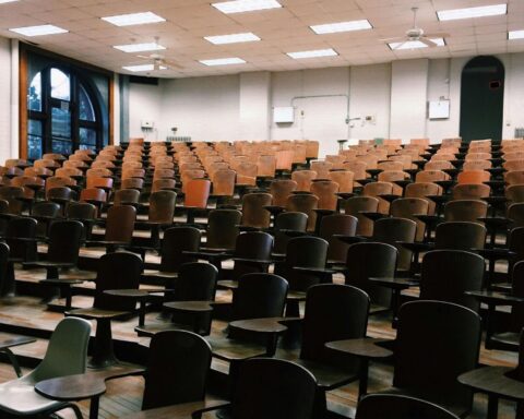 The image depicts an empty school classroom with a ledger of empty seats rowed upwards to doors.