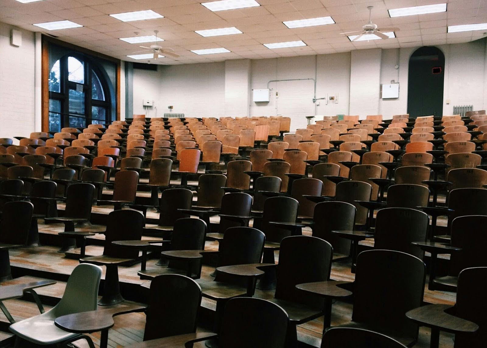 The image depicts an empty school classroom with a ledger of empty seats rowed upwards to doors.