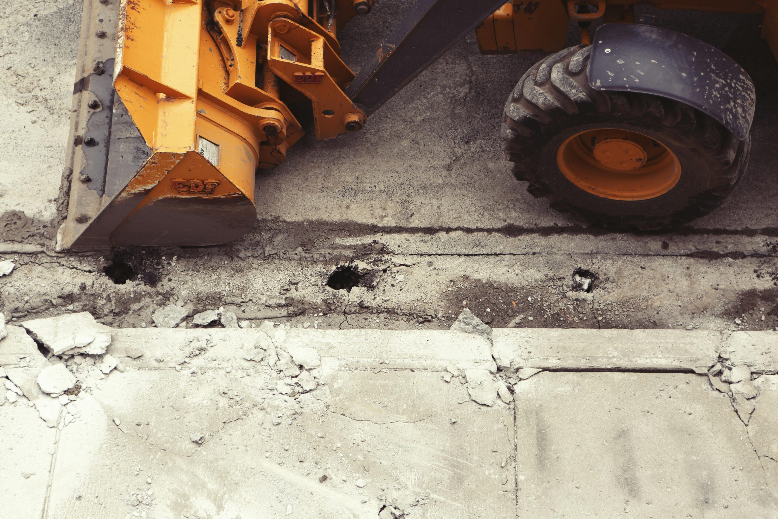 A yellow construction vehicle along a broken, crumbling sidewalk.