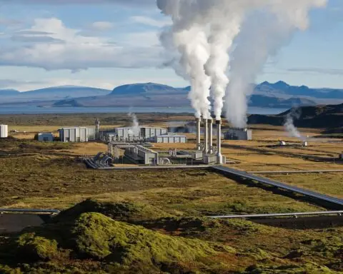 A geothermal power plant as viewed from a hill spewing smoke into the air in a grass valley.