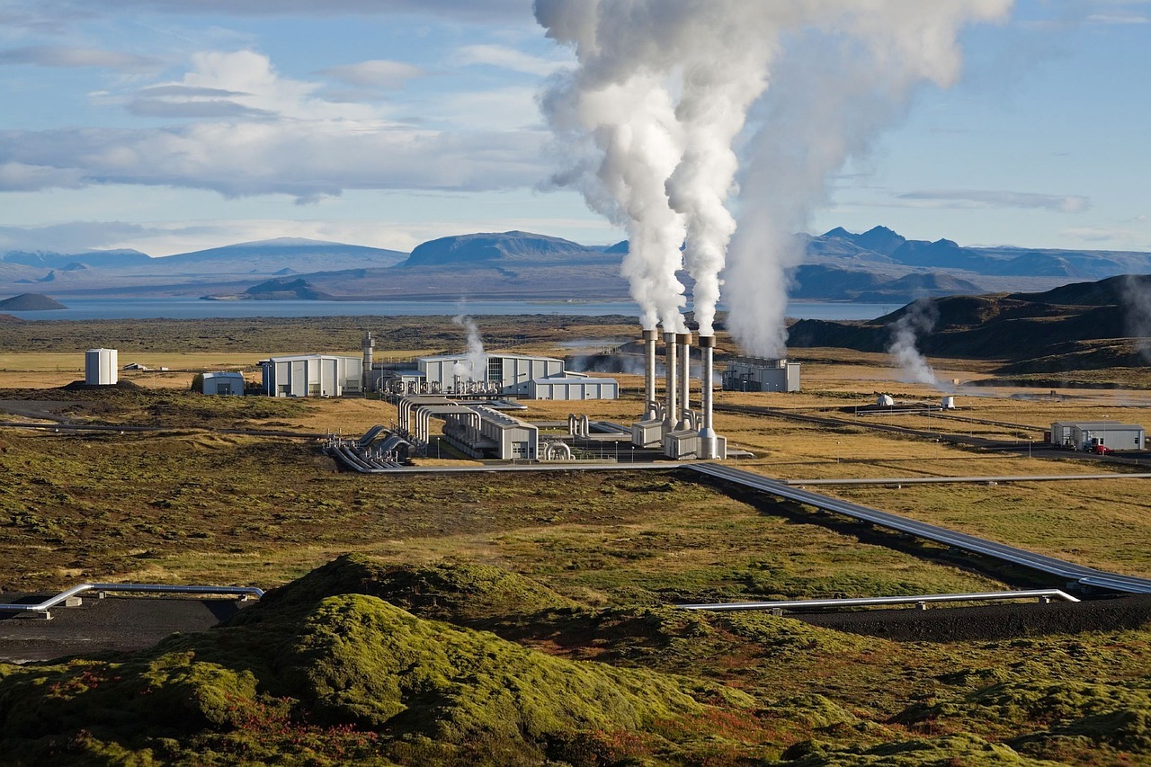 A geothermal power plant as viewed from a hill spewing smoke into the air in a grass valley.