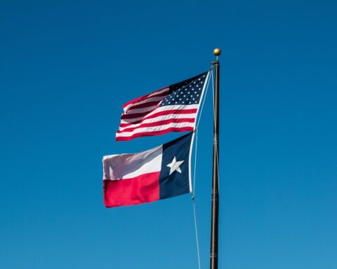 The Texas and United States flags fly together on a flagpole in front of a crisp blue sky.