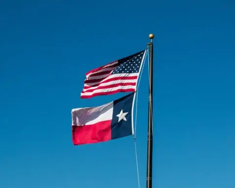 The Texas and United States flags fly together on a flagpole in front of a crisp blue sky.
