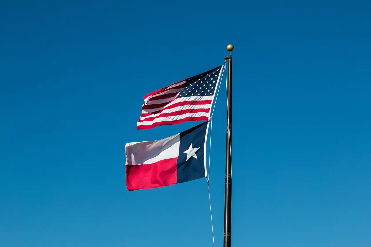 The Texas and United States flags fly together on a flagpole in front of a crisp blue sky.