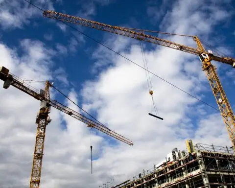 Two cranes are pictured from the ground on a construction site into a partially cloudy sky.