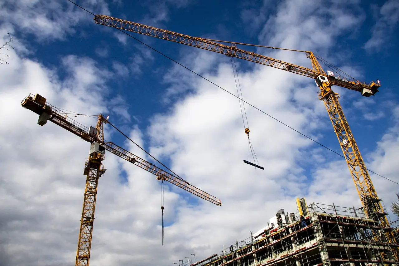 Two cranes are pictured from the ground on a construction site into a partially cloudy sky.