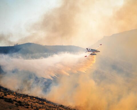 An airplane equipped to fight wildfire appears over flame-engulfed smoke.