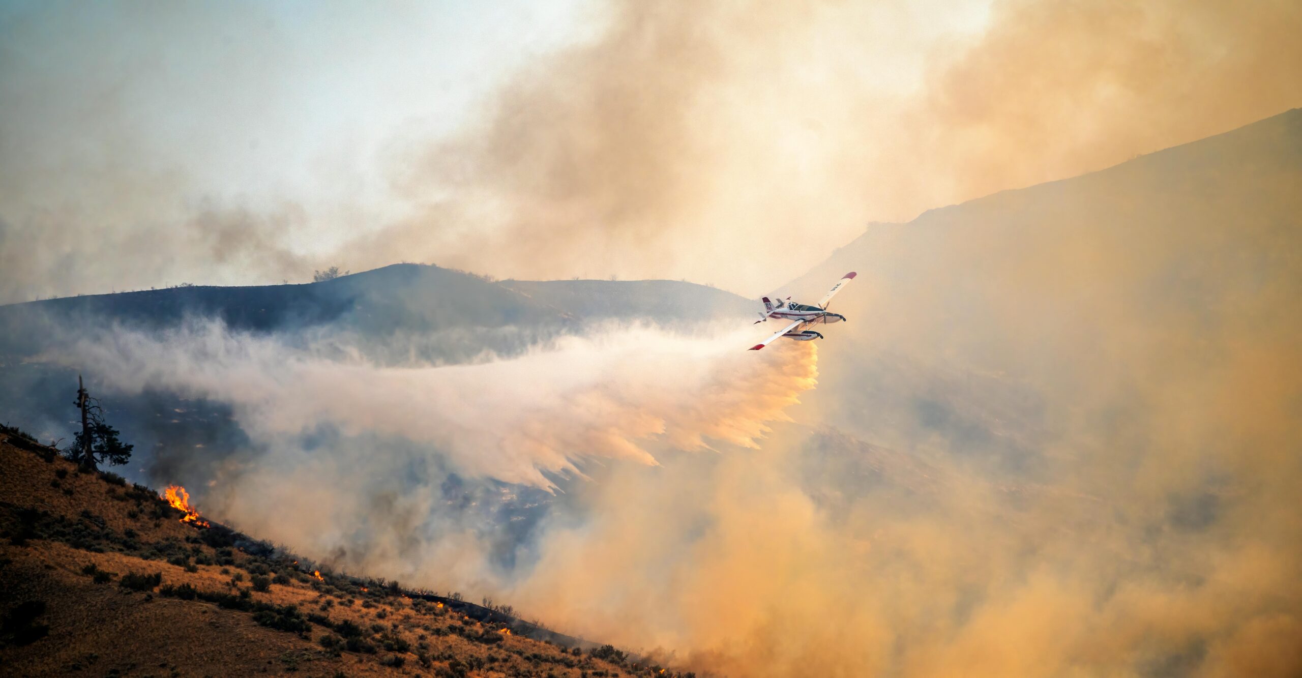 An airplane equipped to fight wildfire appears over flame-engulfed smoke.
