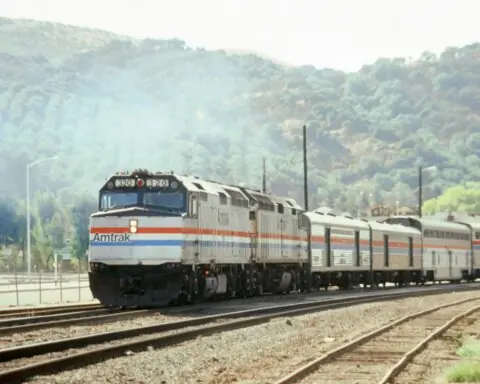 An Amtrak train on a set of tracks before grassy, bushy hills.