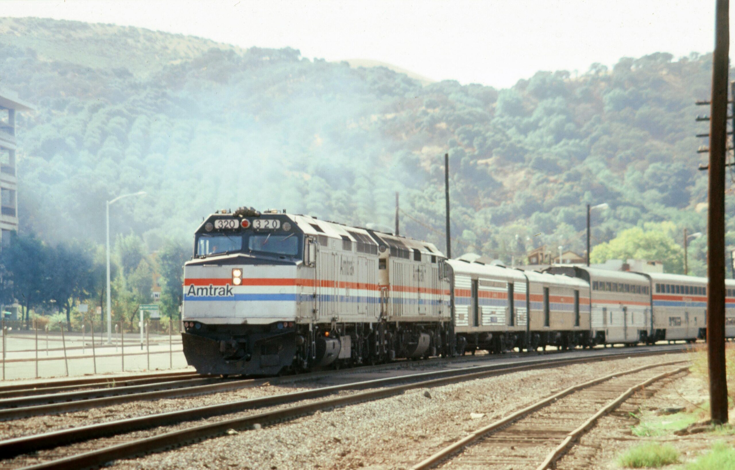 An Amtrak train on a set of tracks before grassy, bushy hills.