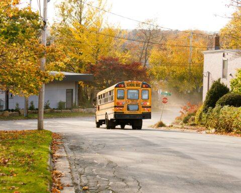 A yellow school bus at a stop sign on a road alongside grass, sidewalks and trees.