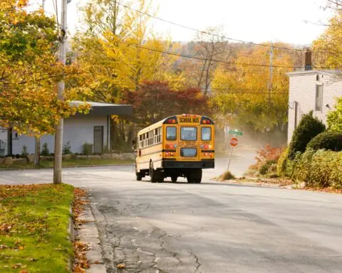 A yellow school bus at a stop sign on a road alongside grass, sidewalks and trees.
