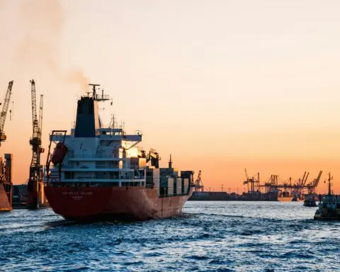 An ocean freightliner transports cargo along a navigable waterway under a setting sun-lit sky.