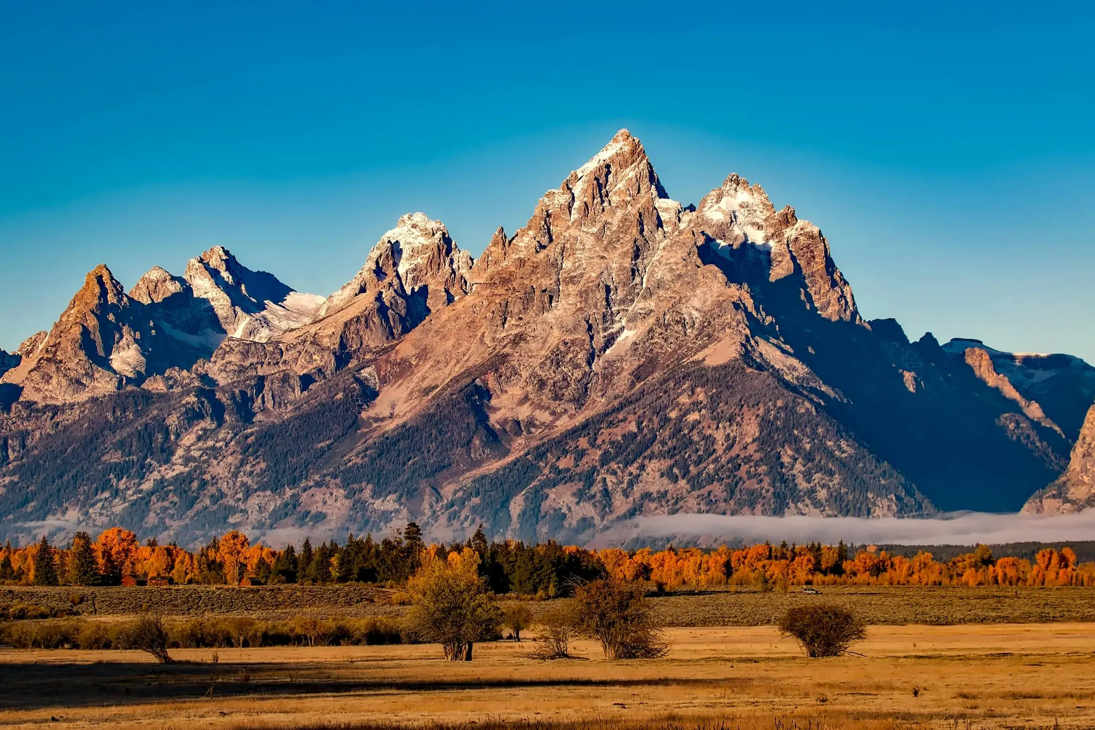 An American national park hosting a mountain range, flat grassy knolls and scattered dry-heat trees.