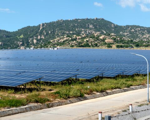 An array of solar panels line a field before a grassy, rocky hill in the background.