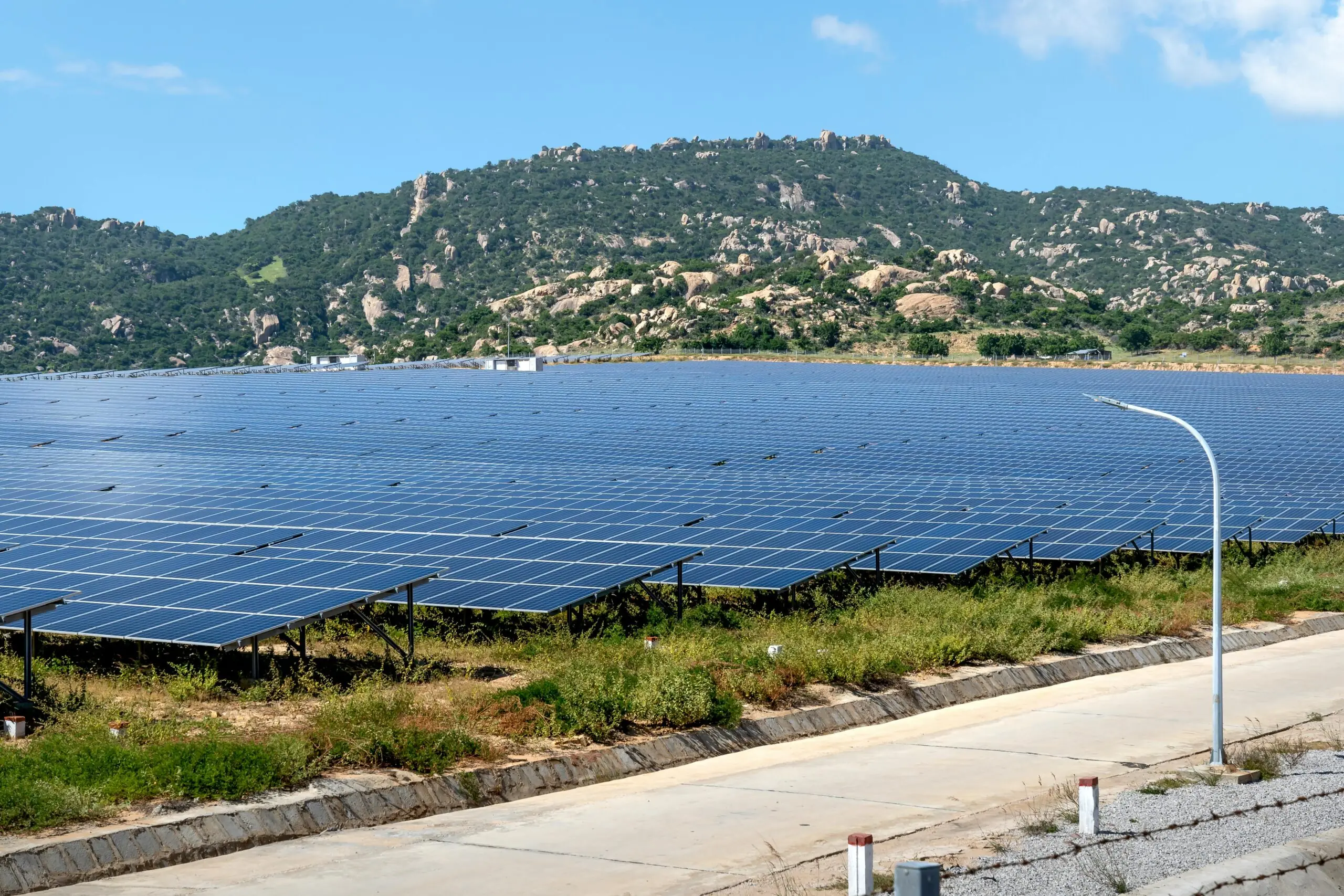 An array of solar panels line a field before a grassy, rocky hill in the background.
