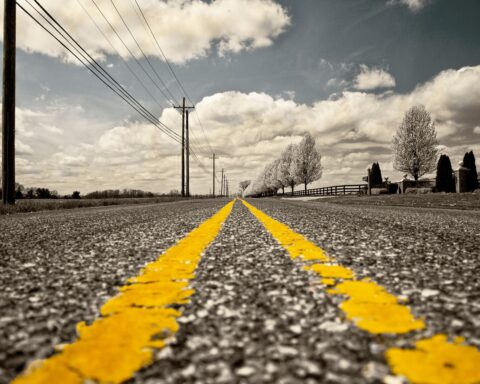 Yellow stripes line an asphalt road in the sun leading to a cloudy blue sky along the horizon.