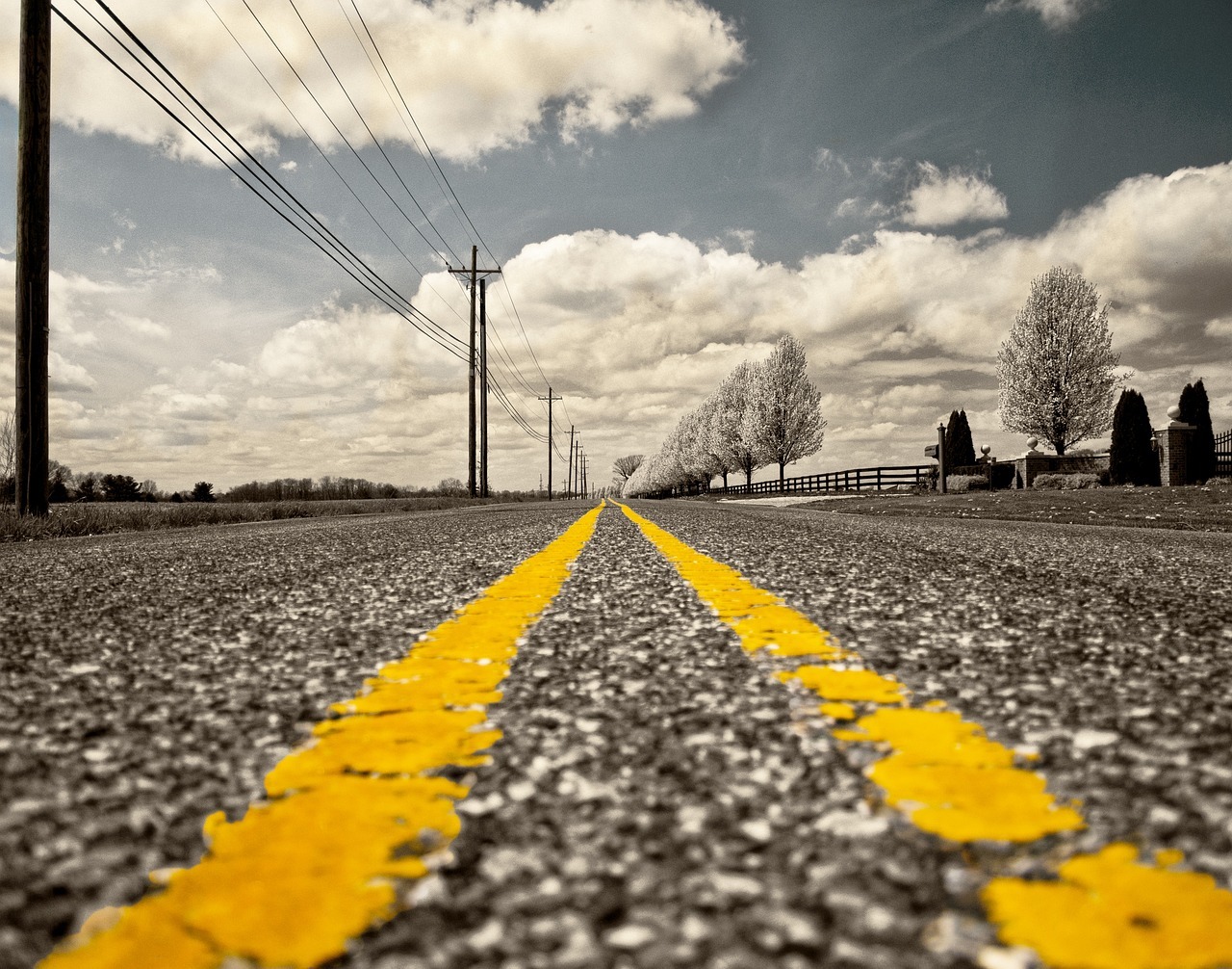 Yellow stripes line an asphalt road in the sun leading to a cloudy blue sky along the horizon.
