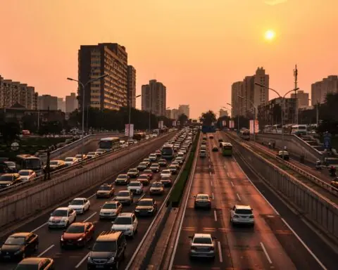 An urban highway packed with cars and trucks is illuminated by a setting sun.