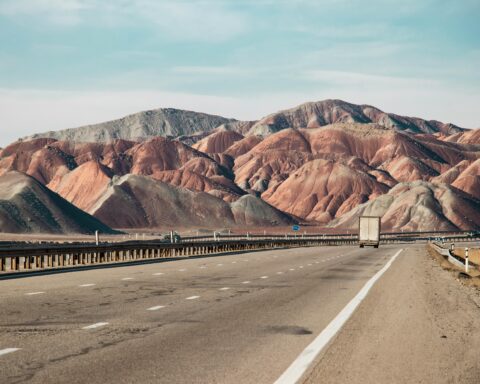 A three-lane highway carrying a semi truck through desert and dry, mountainous country.