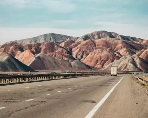 A three-lane highway carrying a semi truck through desert and dry, mountainous country.