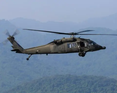 A Blackhawk helicopter in the air above trees and mountainous hills.