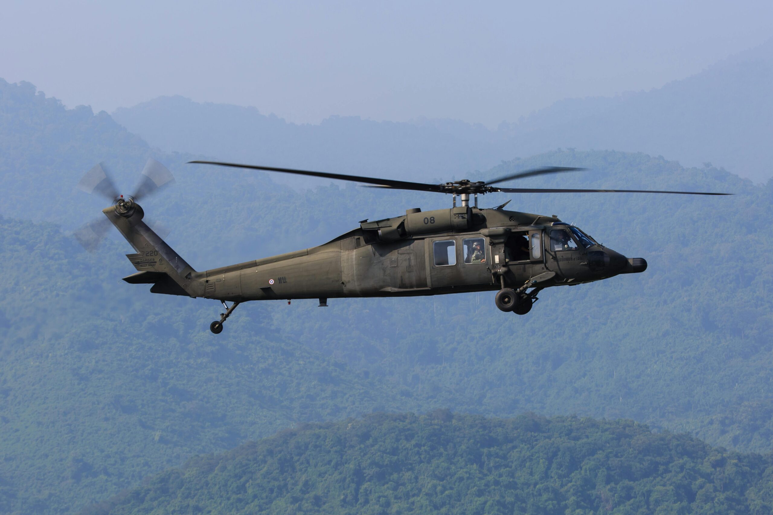 A Blackhawk helicopter in the air above trees and mountainous hills.