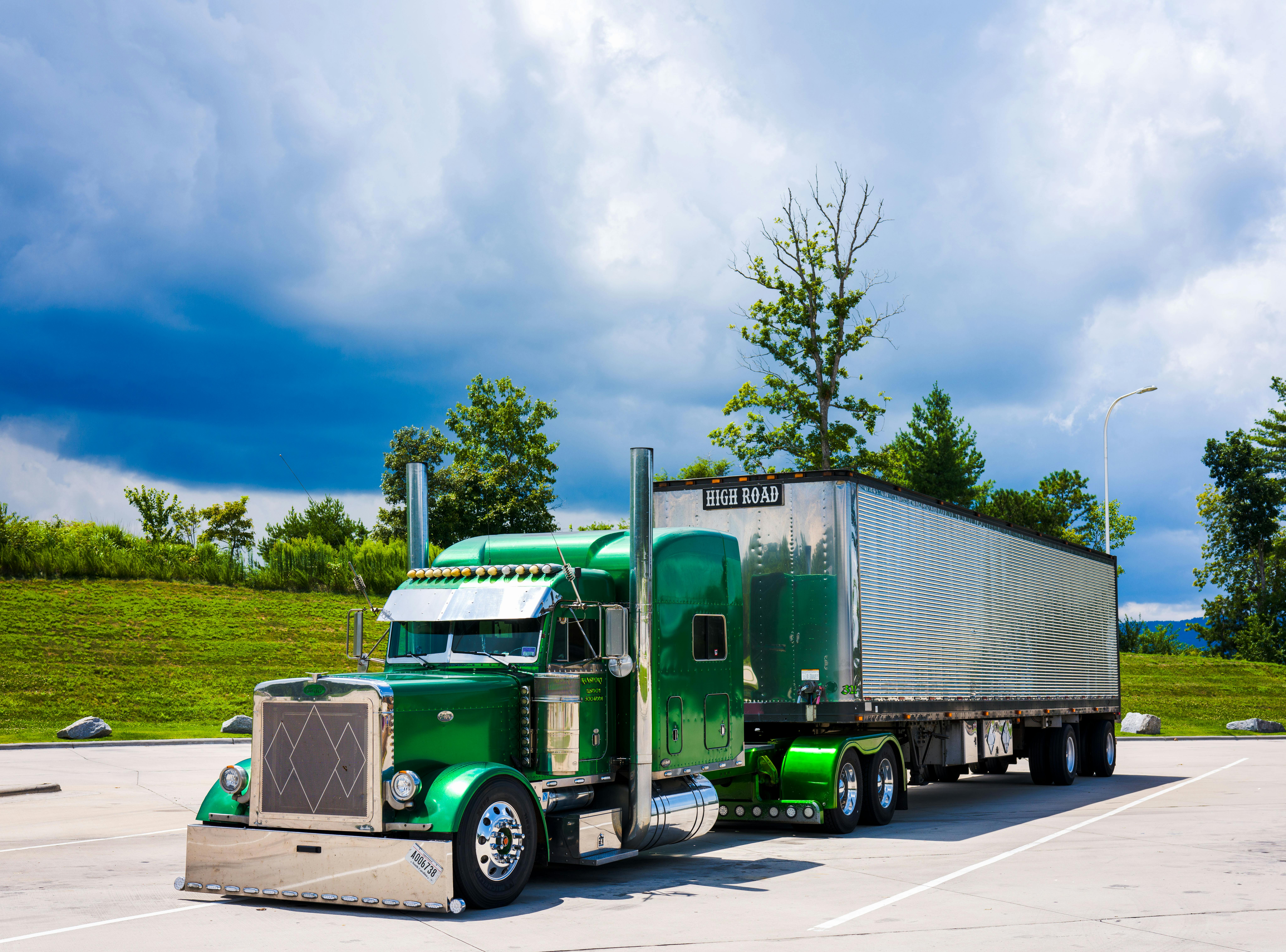 A green semi truck before trees, grass and a cloudy blue sky.