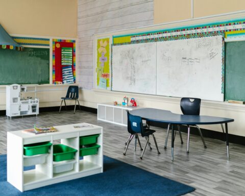 The inside of a school classroom with chairs, desks, small cabinets and storage containers and a chalkboard.