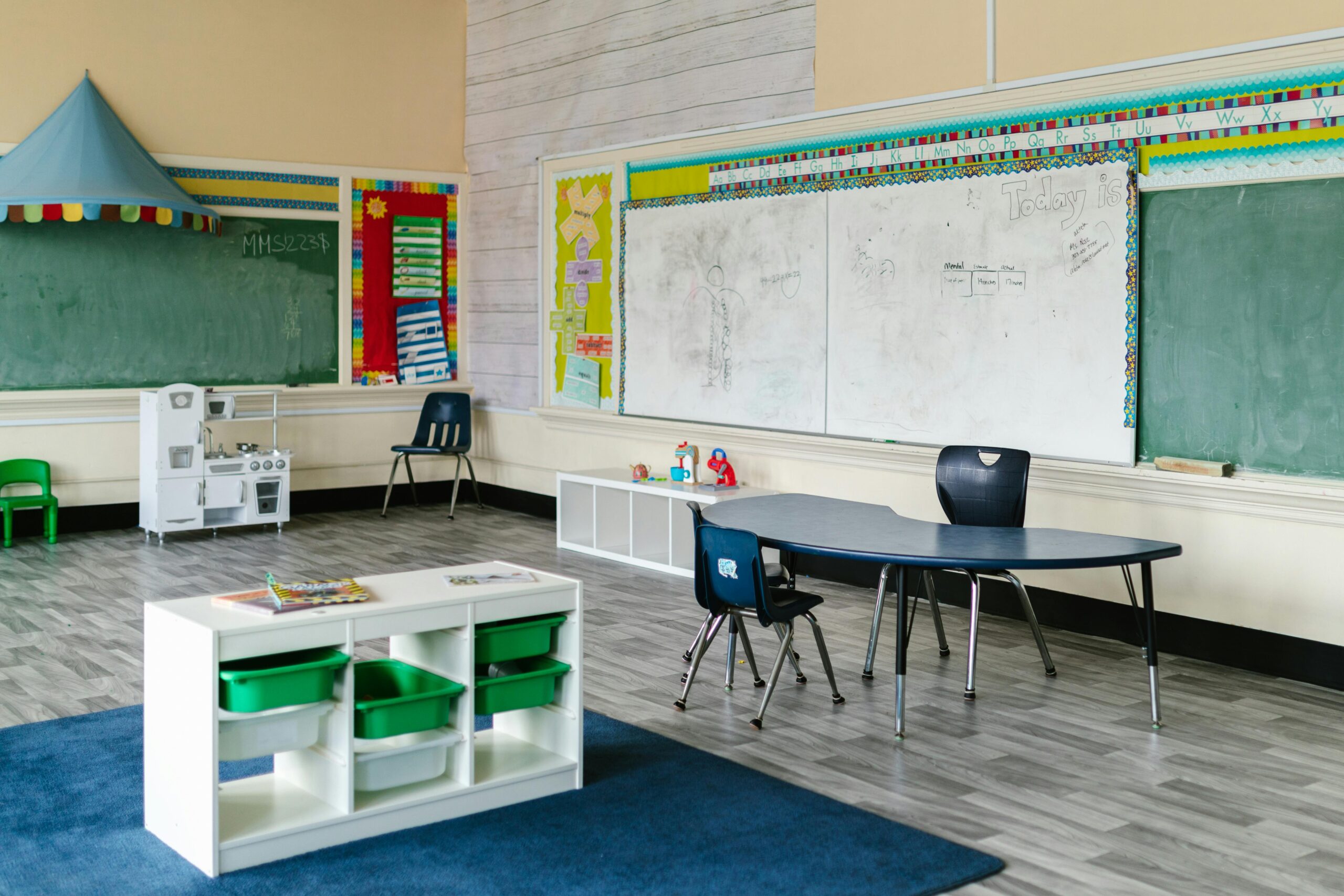 The inside of a school classroom with chairs, desks, small cabinets and storage containers and a chalkboard.