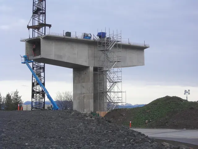 A bridge span under construction before equipment and a cloudy grey sky.