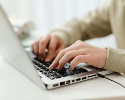 A person types on a silver laptop on a white desk in light.