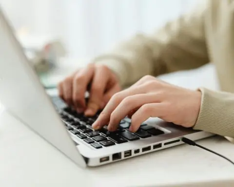 A person types on a silver laptop on a white desk in light.