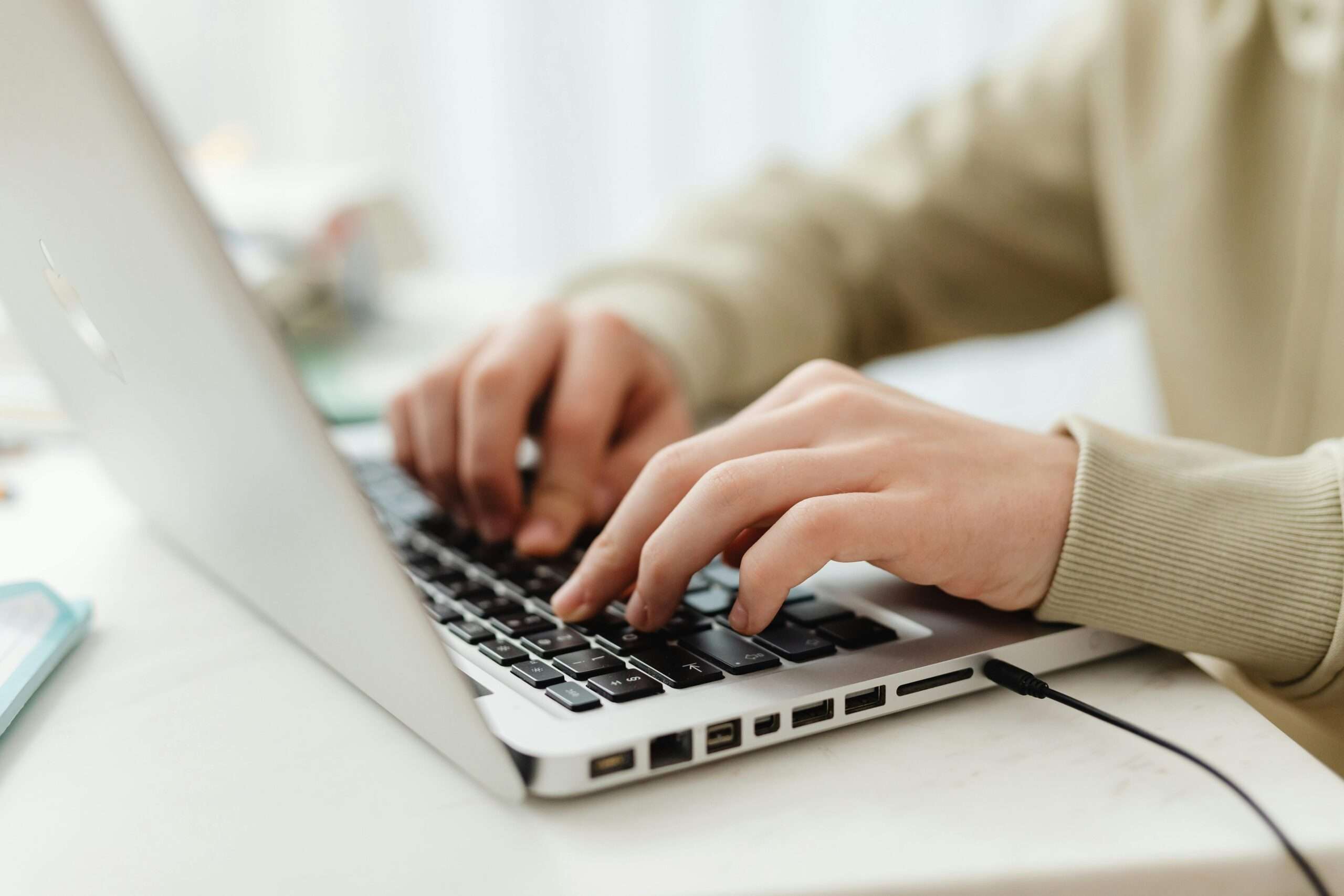 A person types on a silver laptop on a white desk in light.