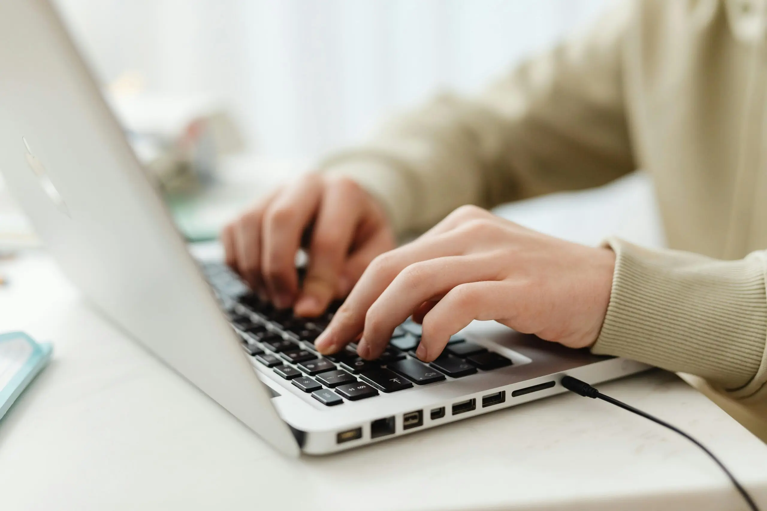 A person types on a silver laptop on a white desk in light.