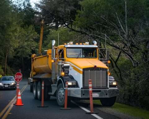 A yellow truck on the side of the road before orange cones and a passing car.