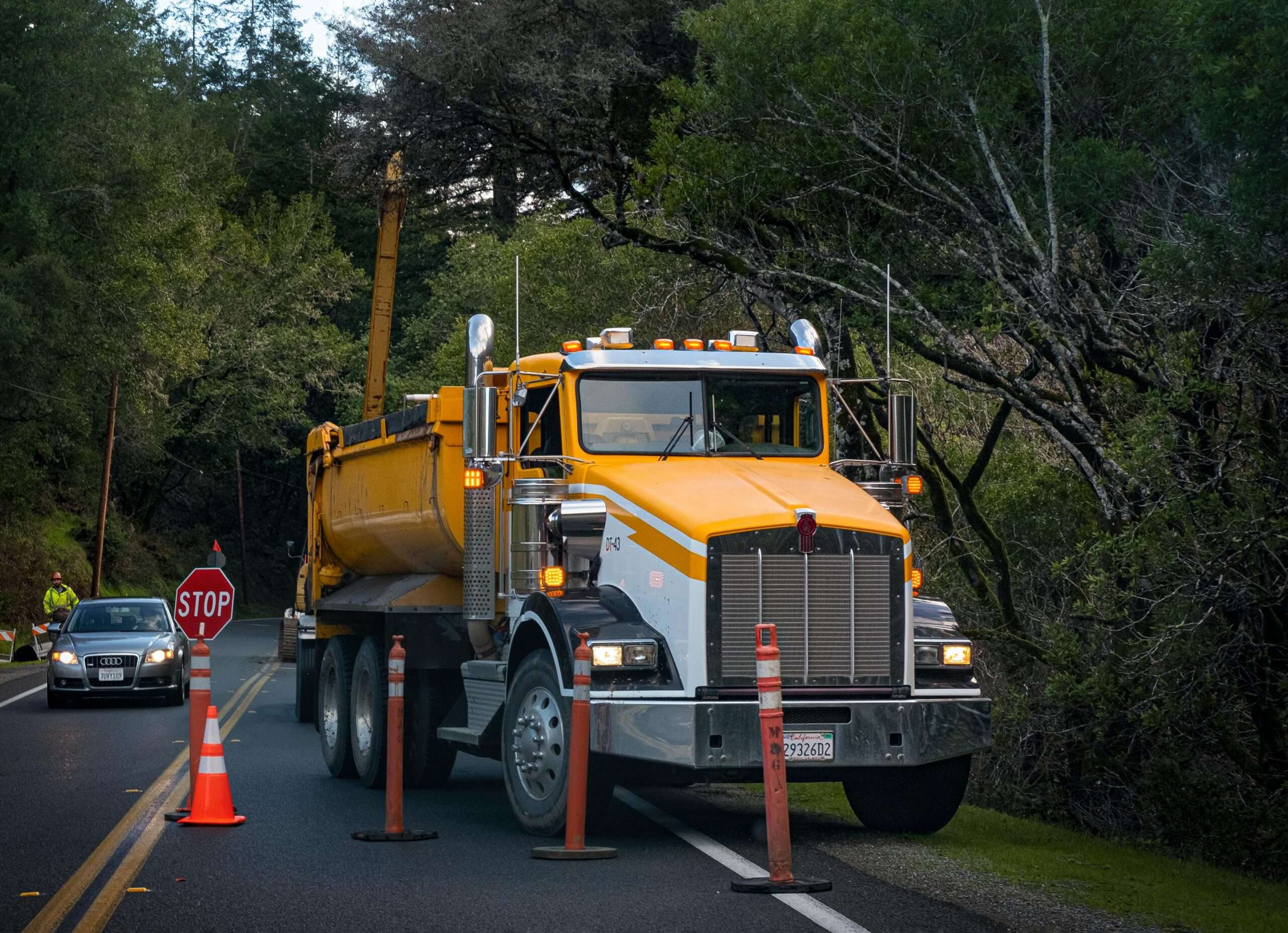 A yellow truck on the side of the road before orange cones and a passing car.