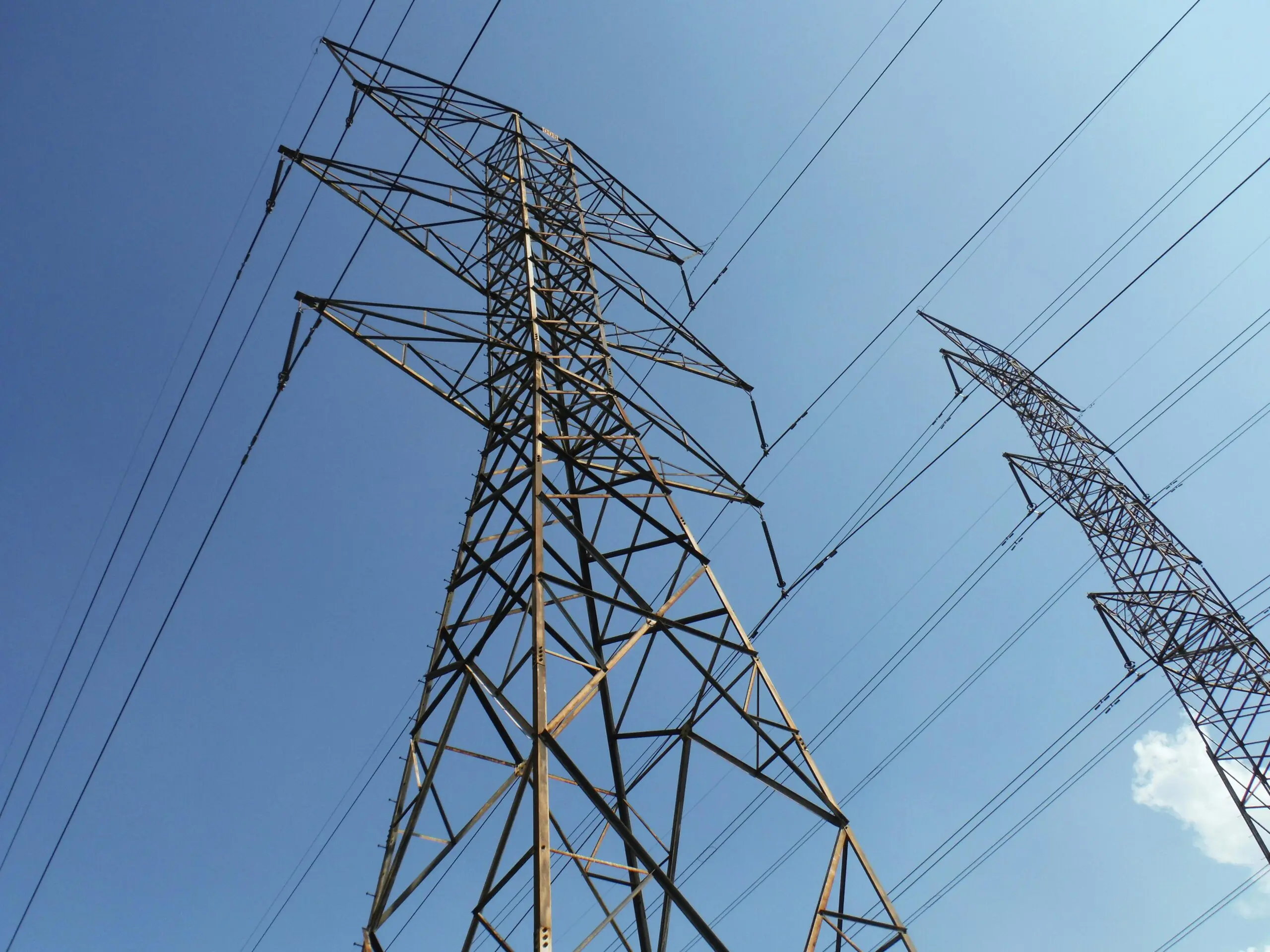 Electric transmission towers before a cloudy blue sky from teh ground.