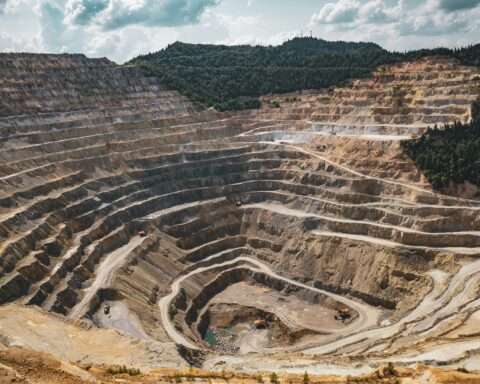 A layered mine with rock and gravel before a forest and a cloudy blue sky.