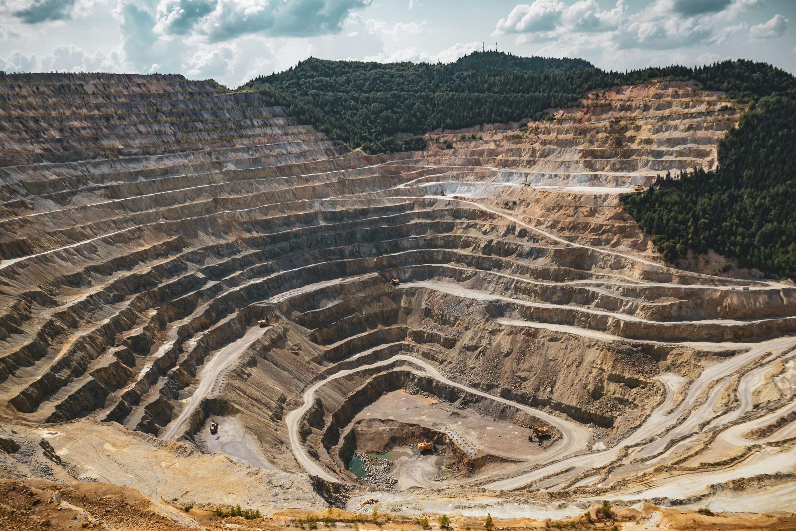 A layered mine with rock and gravel before a forest and a cloudy blue sky.