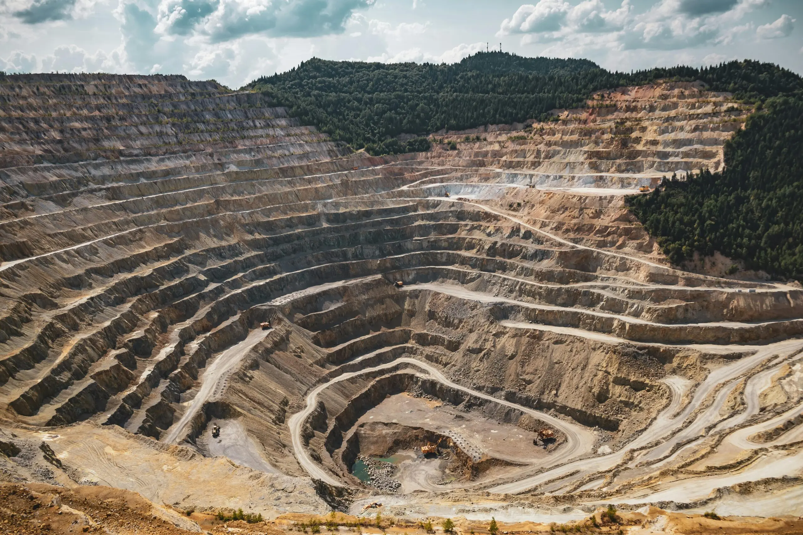 A layered mine with rock and gravel before a forest and a cloudy blue sky.
